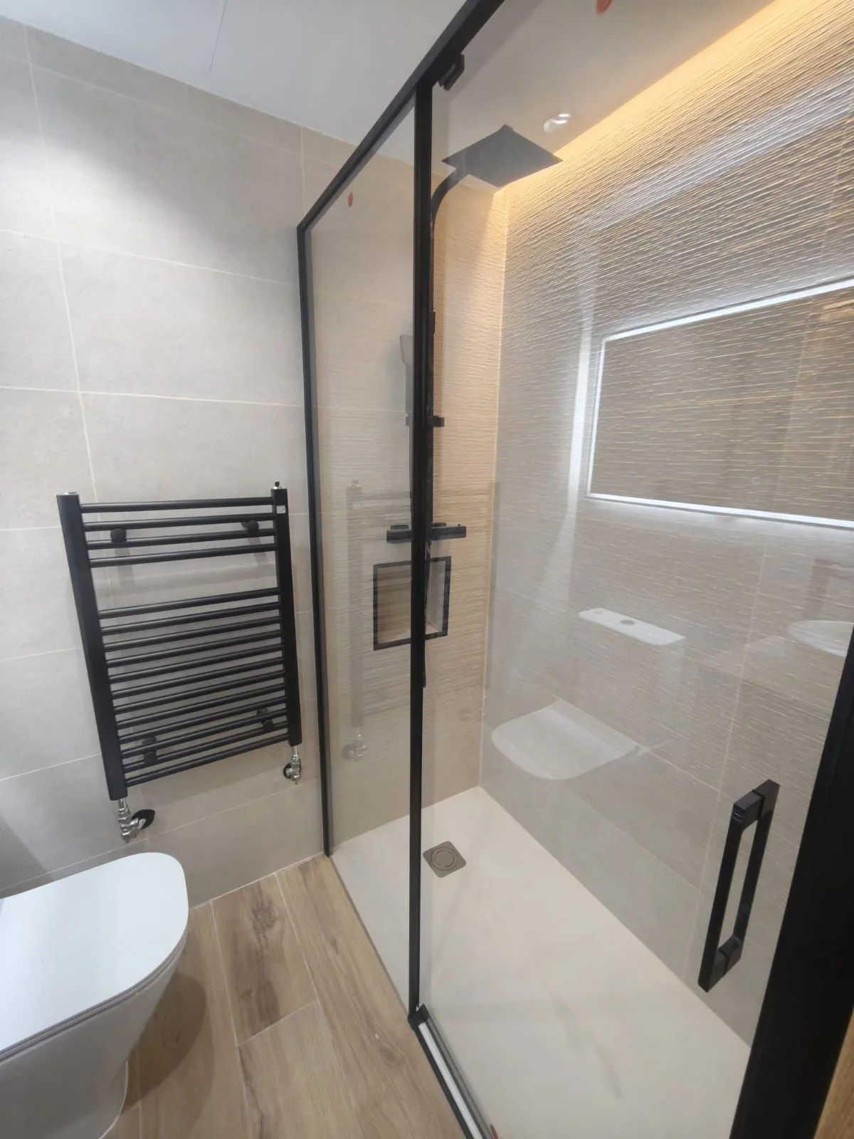Plato de ducha con mampara de cristal y perfiles negros, nicho iluminado y pared con relieve