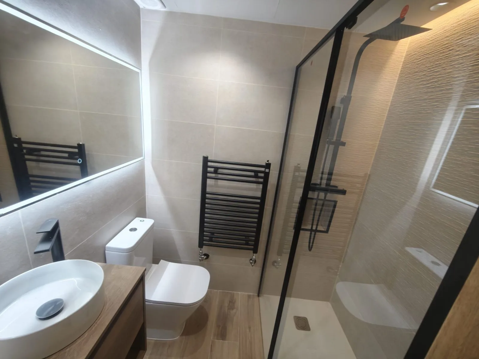Baño reformado con mampara negra, radiador toallero, espejo con luz LED y mueble de madera con lavabo